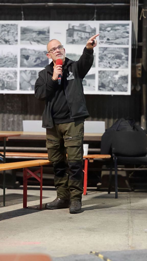 Ein Mann steht in einer Halle, spricht mit einem Mikrofon und zeigt mit dem Finger auf etwas. Im Hintergrund sind mehrere Schwarz-Weiß-Fotos an einer Wand angebracht.