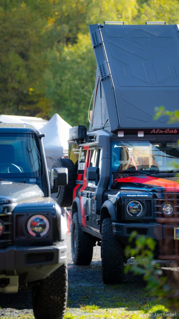 Zwei Geländewagen, einer rot und schwarz mit einem aufklappbaren Dach, stehen auf einem gravel Untergrund, umgeben von Bäumen und einem Zelt im Hintergrund.