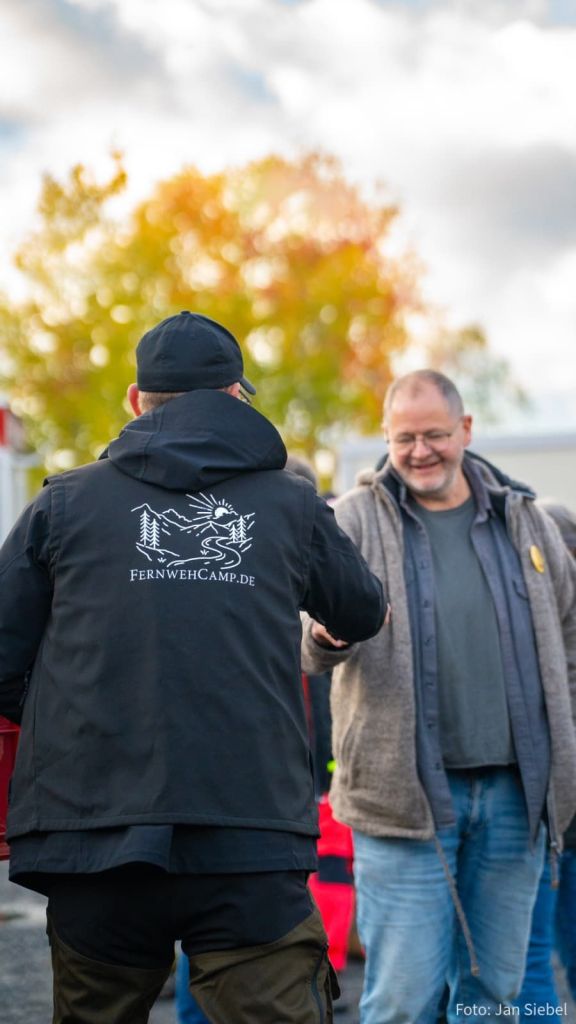 Zwei Männer stehen in einem freundlichen Gespräch, während sie sich die Hände schütteln. Der Mann im Vordergrund trägt eine graue Jacke und lächelt, während der andere Mann einen schwarzen Hoodie mit dem Logo von fernwehcamp.de trägt.