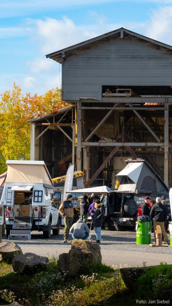 Eine Outdoor-Veranstaltung mit mehreren Offroad-Fahrzeugen und Campern, Personen an einem früheren Industriegebäude, umgeben von bunten Herbstbäumen.