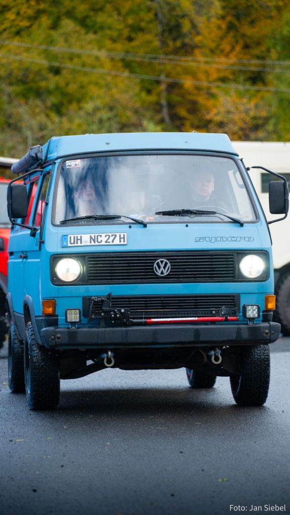 Ein blauer Volkswagen Van fährt auf einer nassen Straße. Im Fahrer- und Beifahrerbereich sind zwei Personen zu sehen.