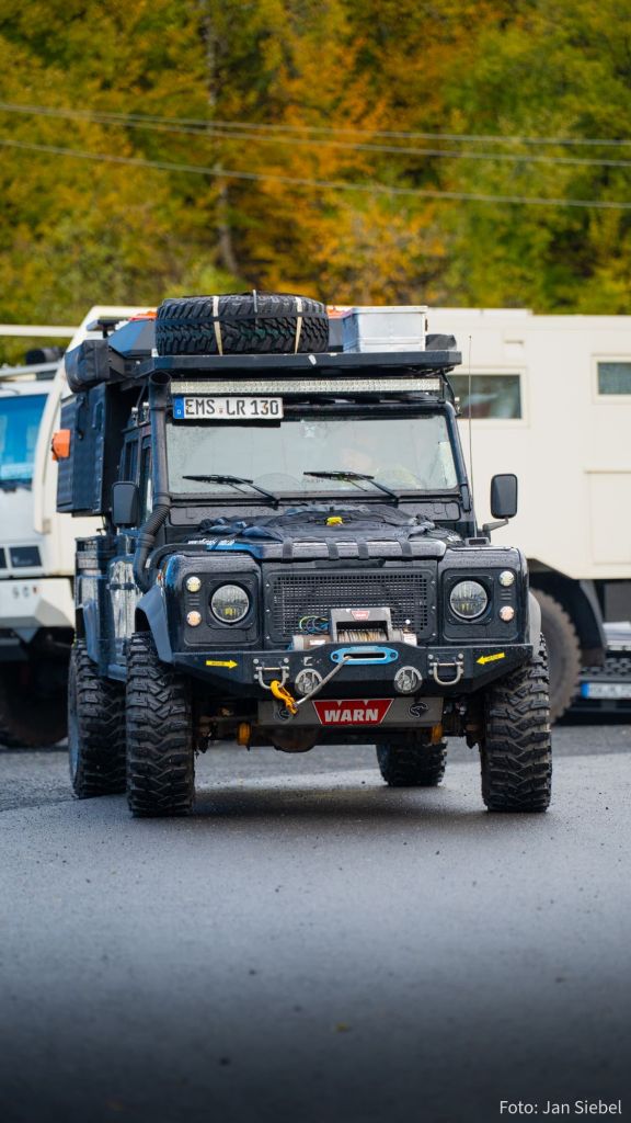 Ein offroad Fahrzeug mit einer robusten Bauweise, großen Reifen und einem Ersatzrad auf dem Dach, fotografiert auf einem Parkplatz mit herbstlicher Baumkulisse im Hintergrund.