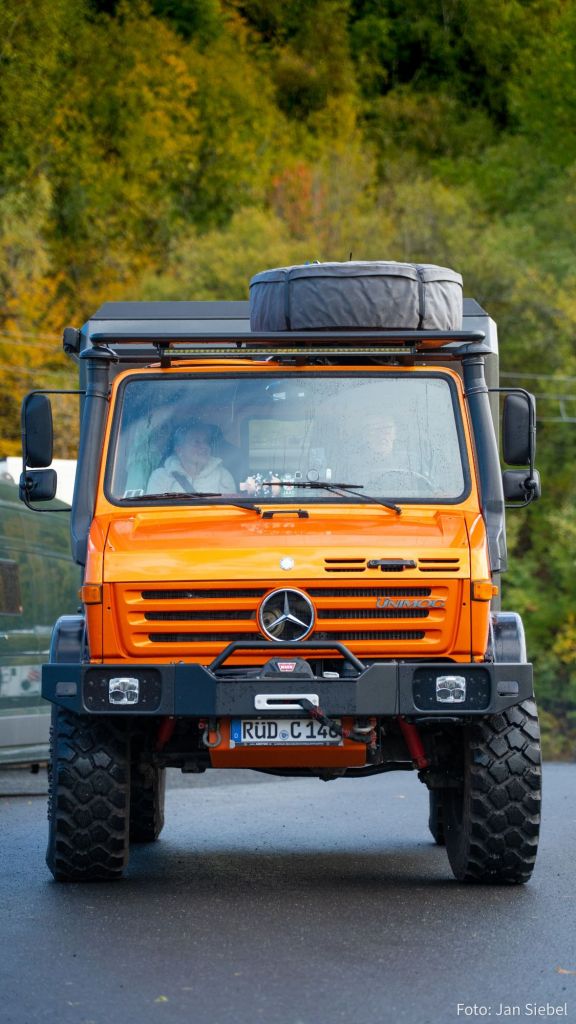 Ein orangefarbener Mercedes Unimog mit einem Ersatzreifen auf dem Dach fährt auf einer Straße, umgeben von einer farbenfrohen herbstlichen Landschaft.